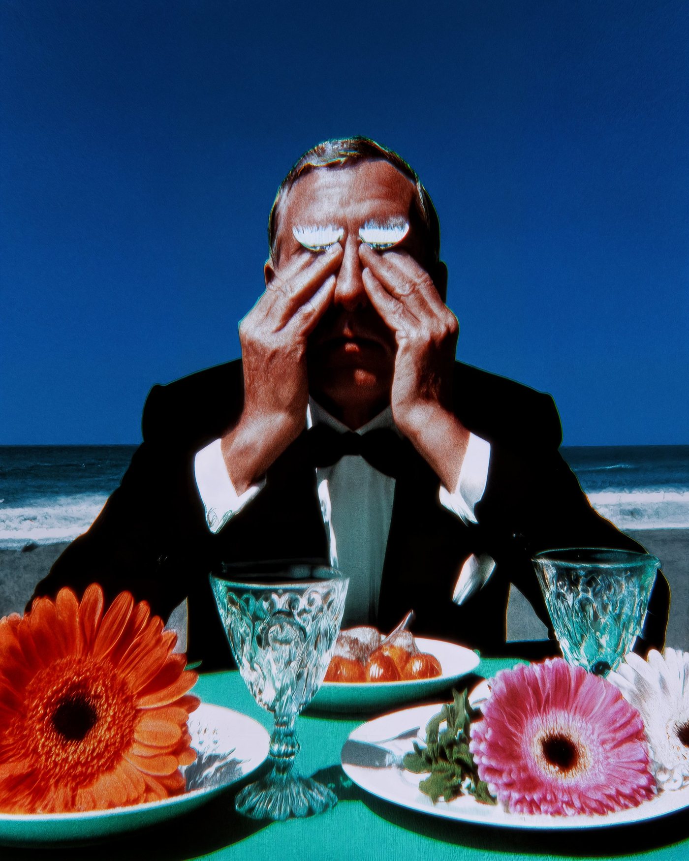 Man in a tuxedo covering his eyes at a table set with gerbera flowers and crystal glasses by the sea